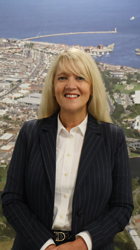 head-and-shoulders portrait of a person wearing a navy pinstripe blazer and white shirt, standing in front of a large aerial photograph of a coastal city and harbour. The person is facing the camera with hands clasped in front, with the coastline, sea and marina visible in the background.