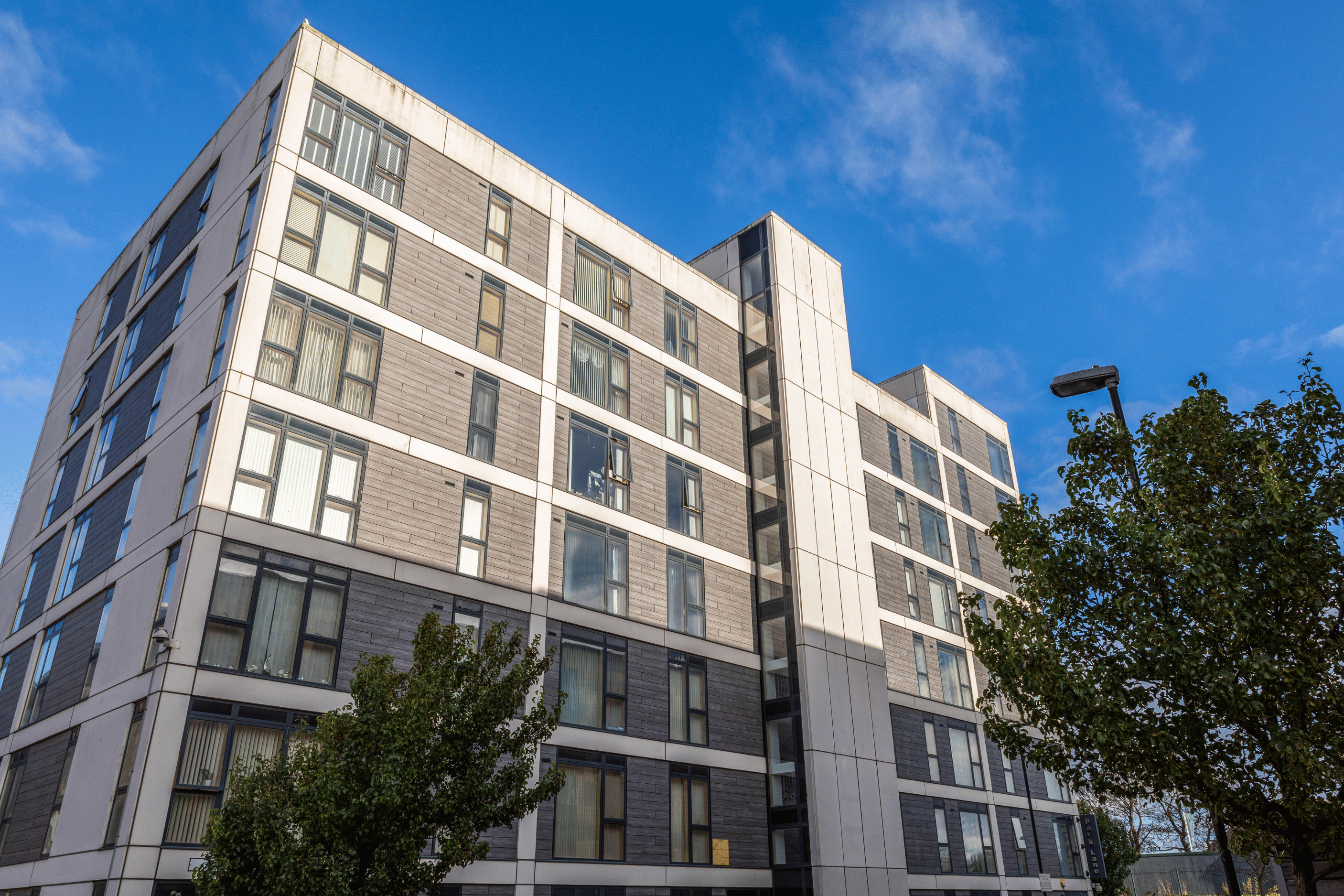 Exterior of a modern multi-storey apartment building with large windows, trees in the foreground, and a blue sky.