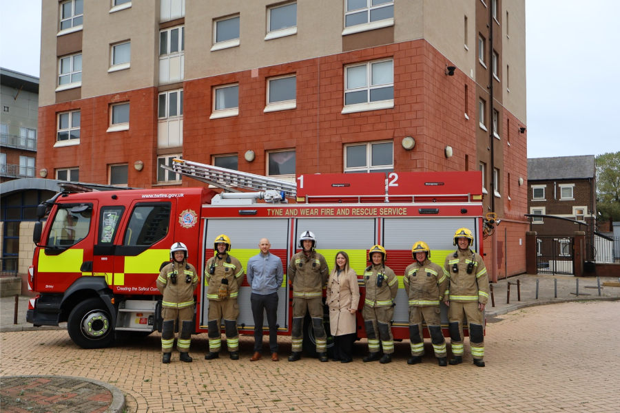 A group of eight people standing in front of a red Tyne and Wear Fire and Rescue Service fire engine, with a multi-storey residential building in the background.